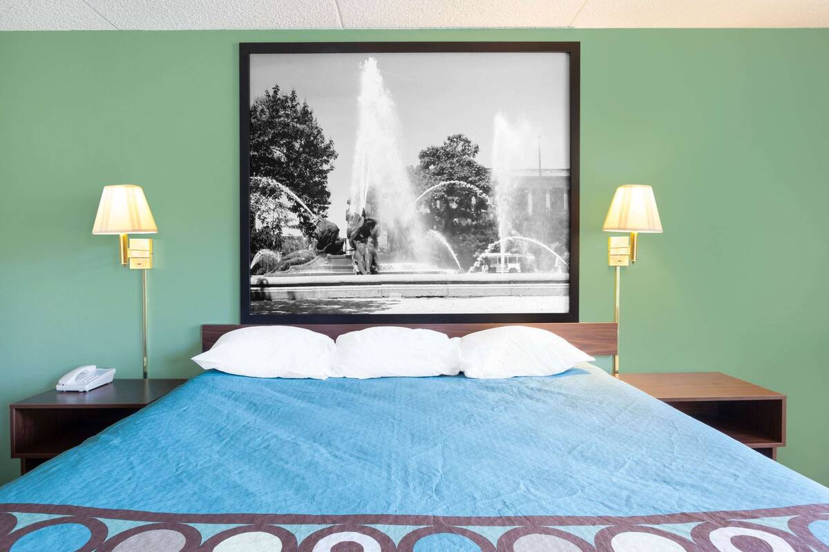 A hotel room with a bed, two bedside lamps, a framed black and white photo of a fountain, and a telephone on the nightstand.