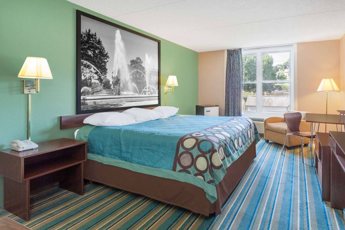 A hotel room with a bed, two bedside lamps, a desk, a chair, a window, and a framed picture on the wall.