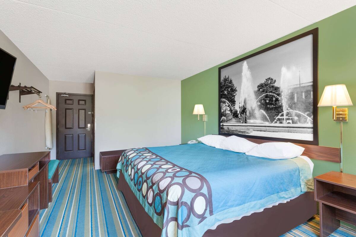 A hotel room with a bed, a desk, a chair, a hanging coat rack, and a large window with a black and white photograph of a fountain.