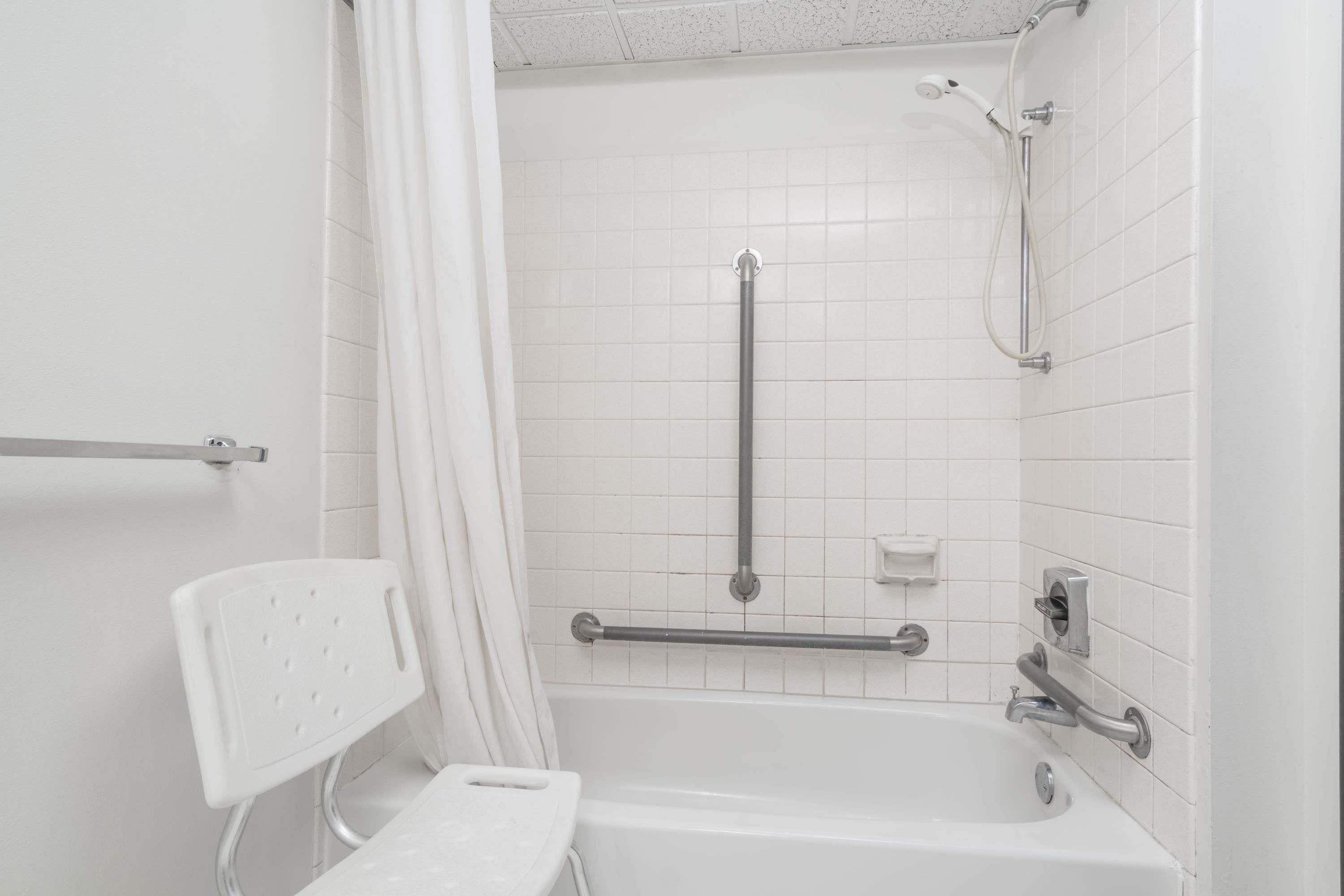 A bathroom with white tiled walls, a white bathtub, a shower with a handheld showerhead, and a grab bar.