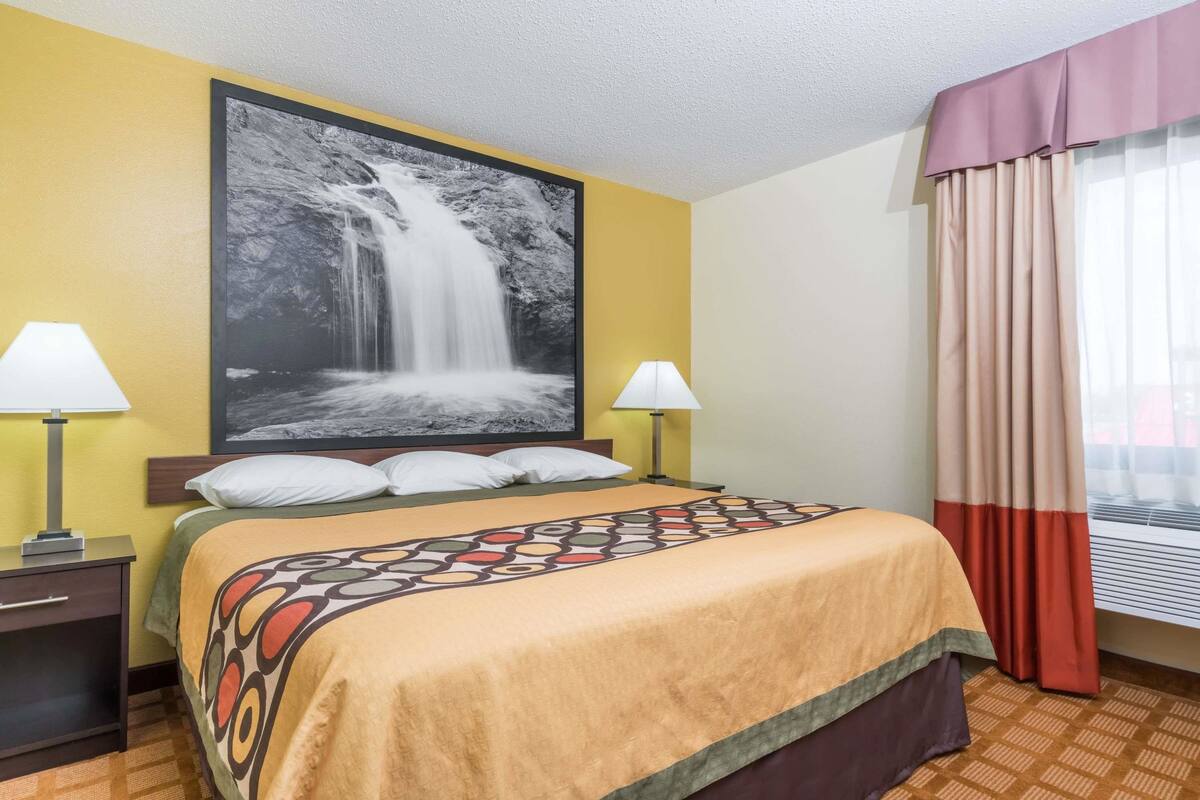 A hotel room with a bed, two bedside lamps, a large waterfall painting on the wall, and a window with curtains.
