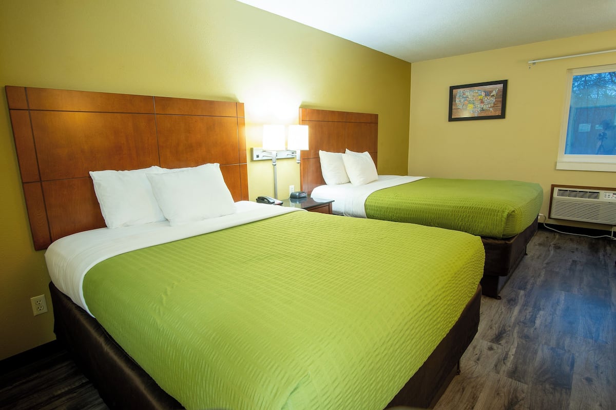 A hotel room with two beds, each with a green bedspread, wooden headboards, and a nightstand with a lamp.
