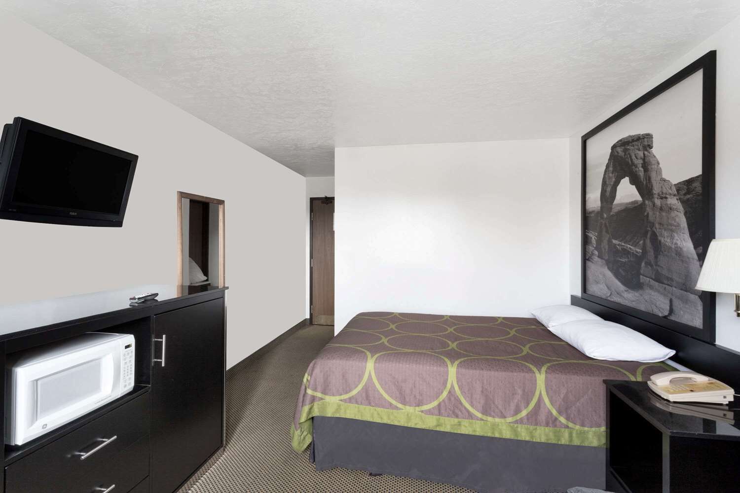 A hotel room with a bed, a television mounted on the wall, a microwave, a desk with a telephone, and a framed picture on the wall.