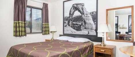 Una camera d'albergo con un letto, un comodino, un tavolo, una finestra con tende, uno specchio fissato al muro e un quadro grande con una formazione rocciosa.