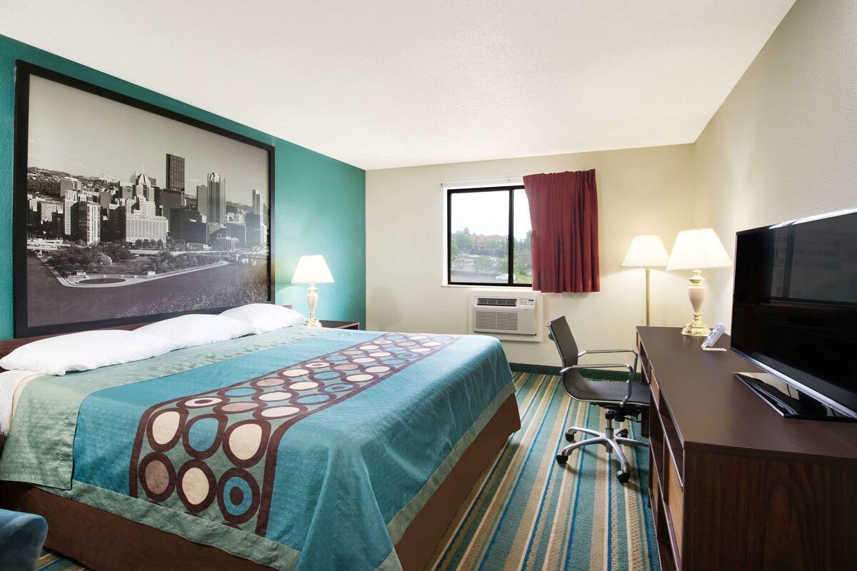 A hotel room with a bed, desk, TV, and a large cityscape painting on the wall.