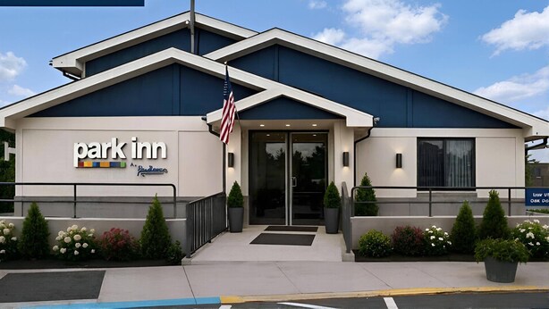 Um hotel Park Inn com design moderno, exibindo uma bandeira americana em destaque, uma entrada acolhedora e paisagismo bem cuidado.