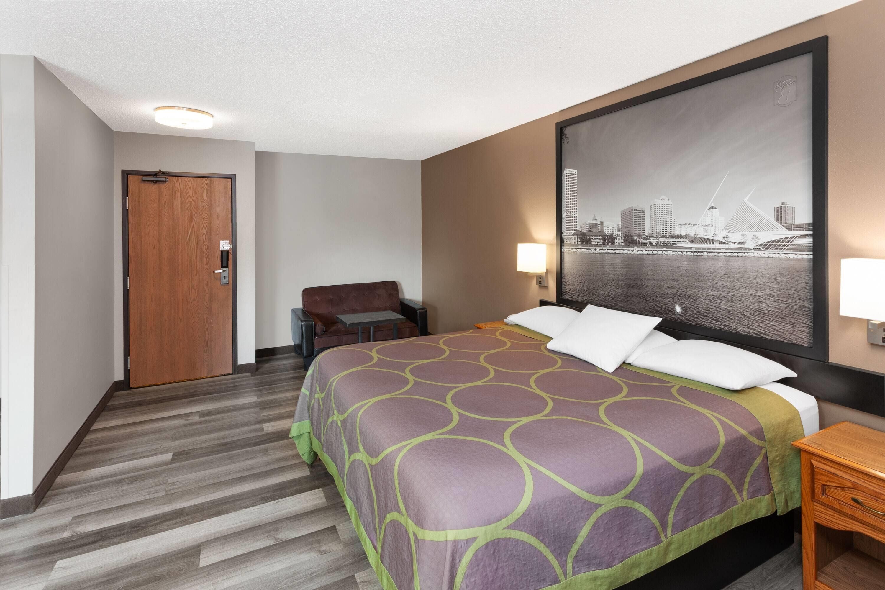 A hotel room with a bed, a chair, a desk, a door, and a cityscape painting on the wall.