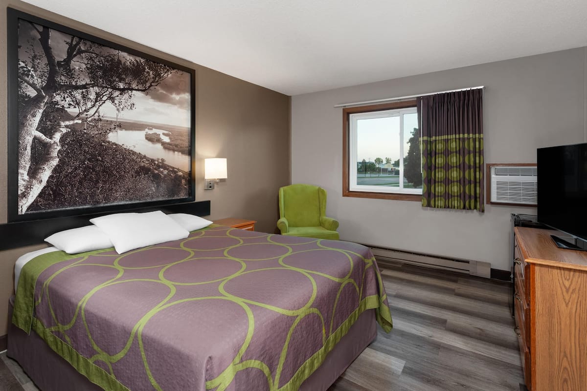Una habitación de hotel con una cama grande, un sillón verde, un televisor y una ventana con cortinas.