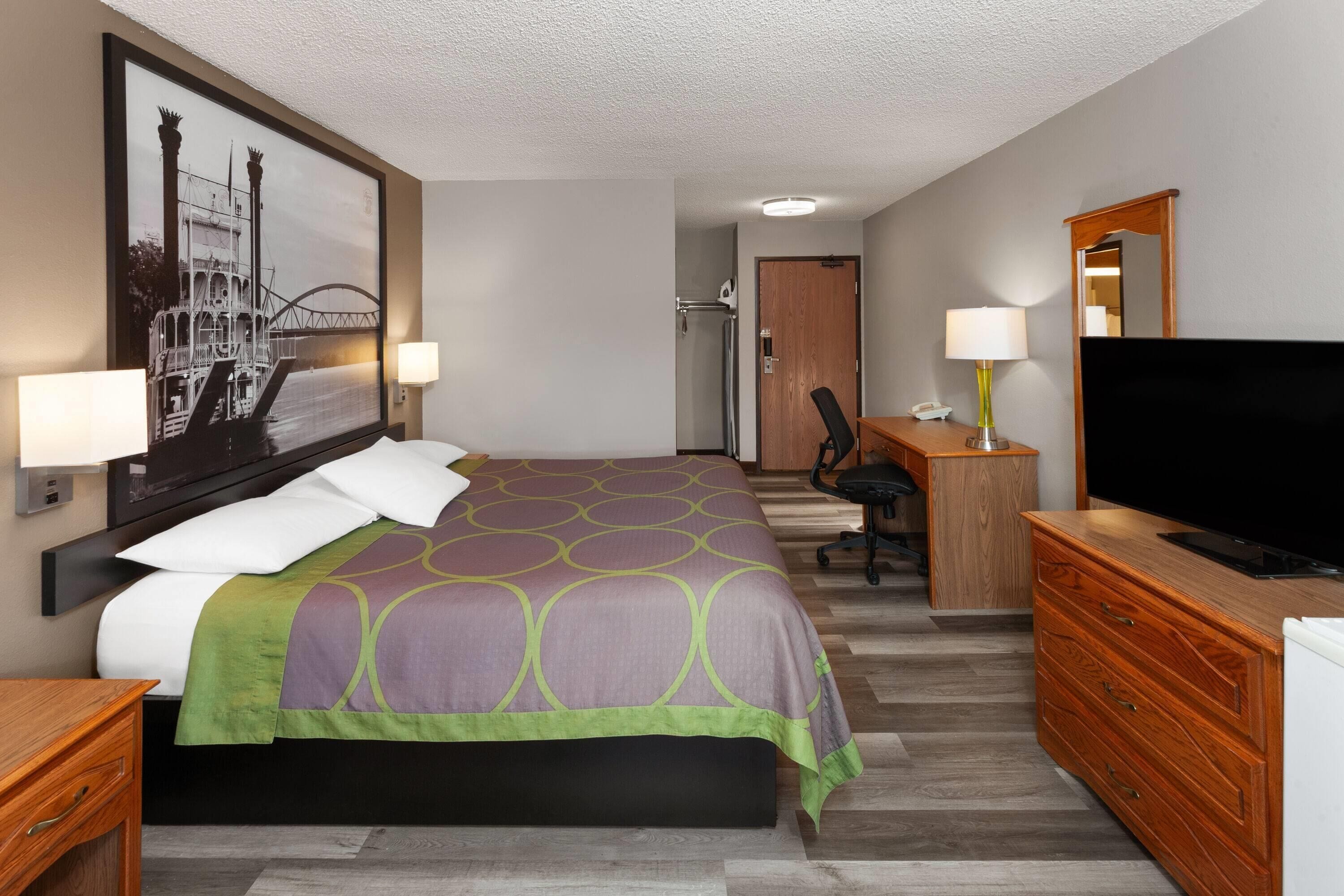 A hotel room with a bed, a desk with a TV, a chair, a dresser, and a painting on the wall.