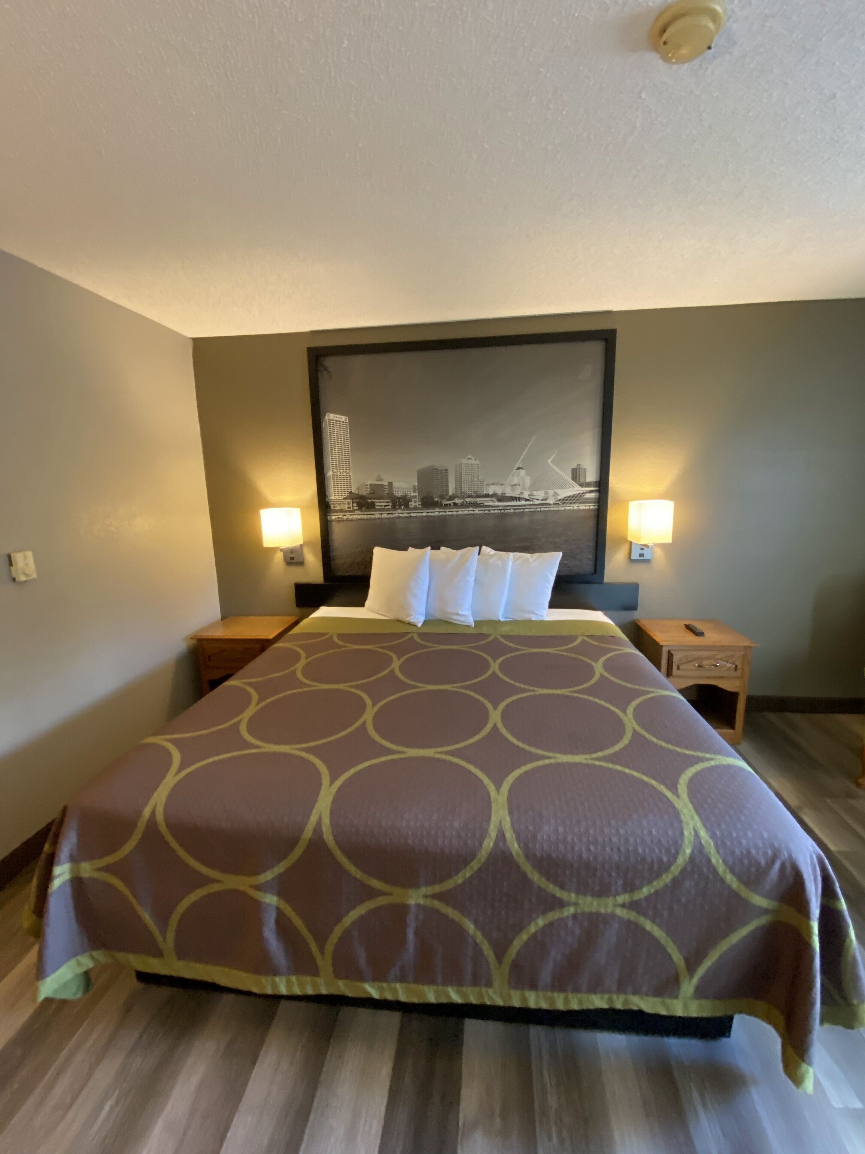 A hotel room with a large bed, two bedside tables, a nightstand, a wall-mounted lamp, and a cityscape painting above the bed.