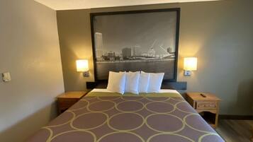 A hotel room with a large bed, two bedside tables, a nightstand, a wall-mounted lamp, and a cityscape painting above the bed.