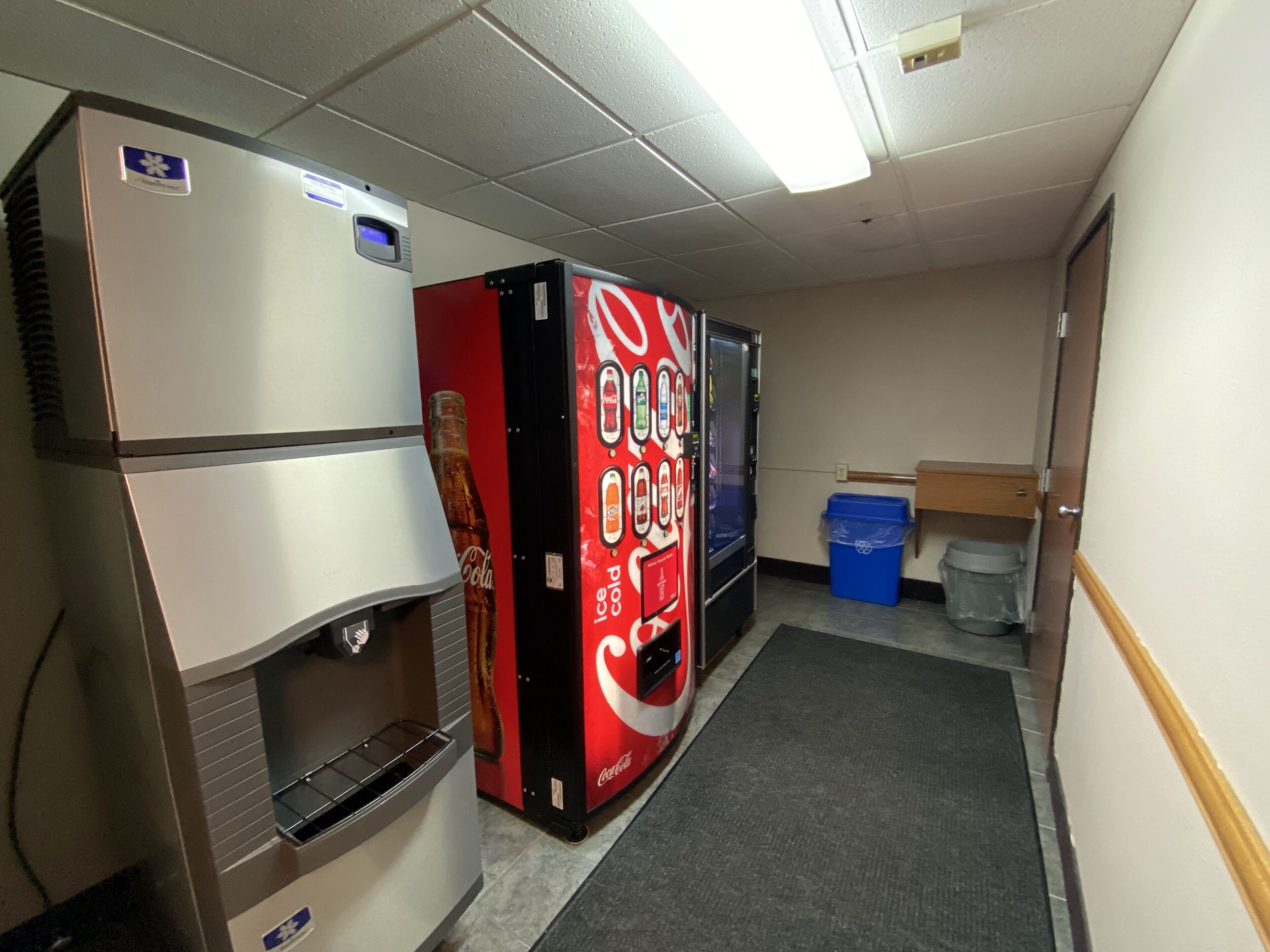 Un pasillo con máquinas expendedoras, incluyendo un refrigerador de Coca-Cola, y un cubo de basura.