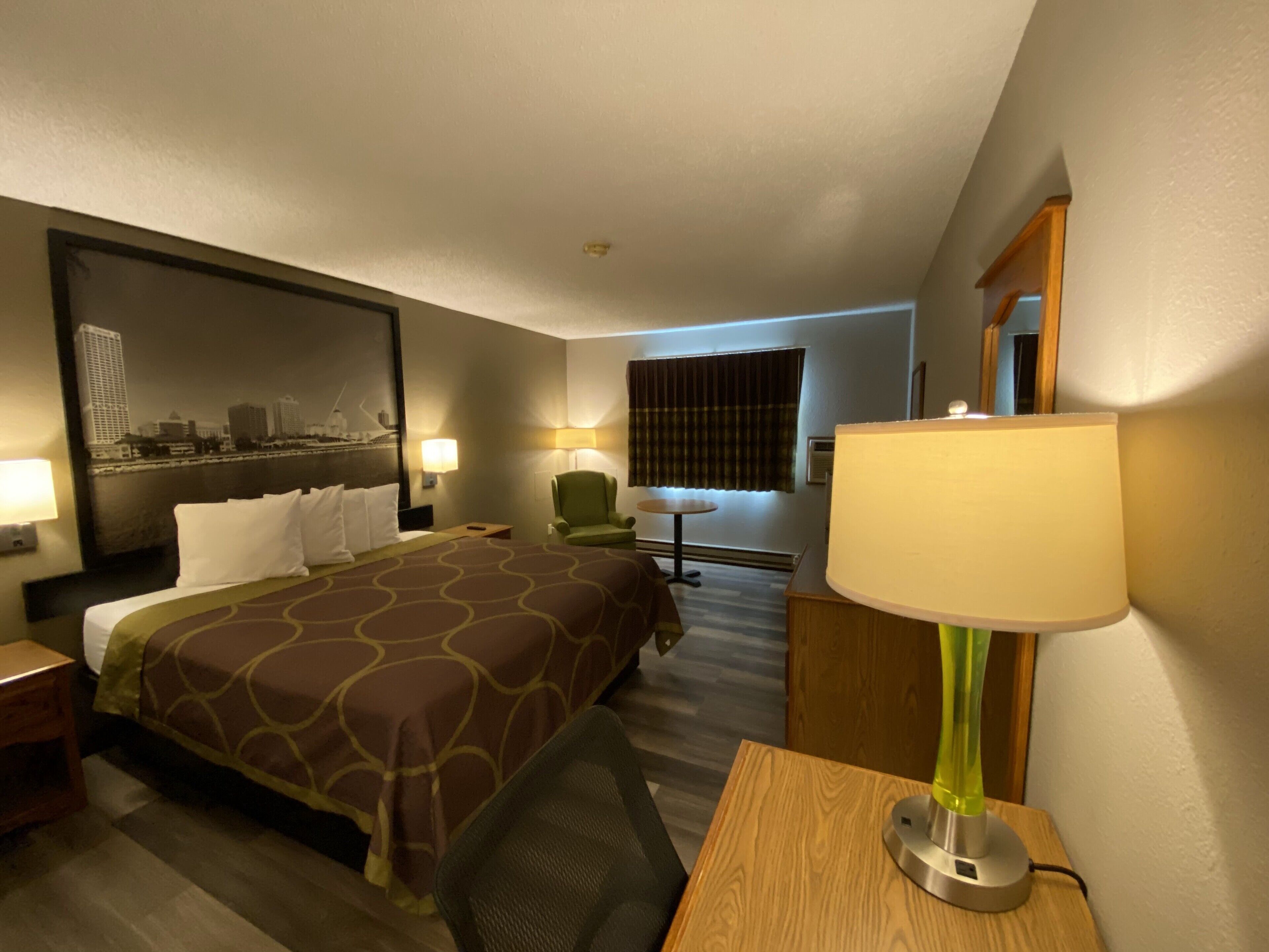 A hotel room with a large bed, a desk, a chair, a lamp, and a view of a cityscape through the window.