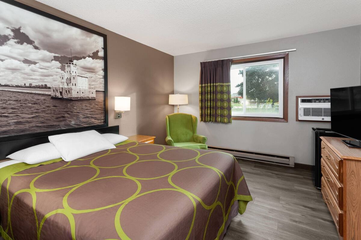 Habitación de hotel con una cama grande, una silla verde, un televisor y una ventana con cortinas.