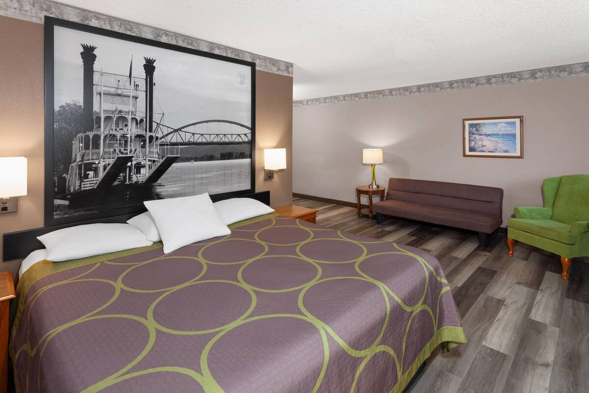 Una habitación de hotel con una cama grande, un sofá, un sillón verde, un cuadro en la pared y una lámpara en la mesita de noche.