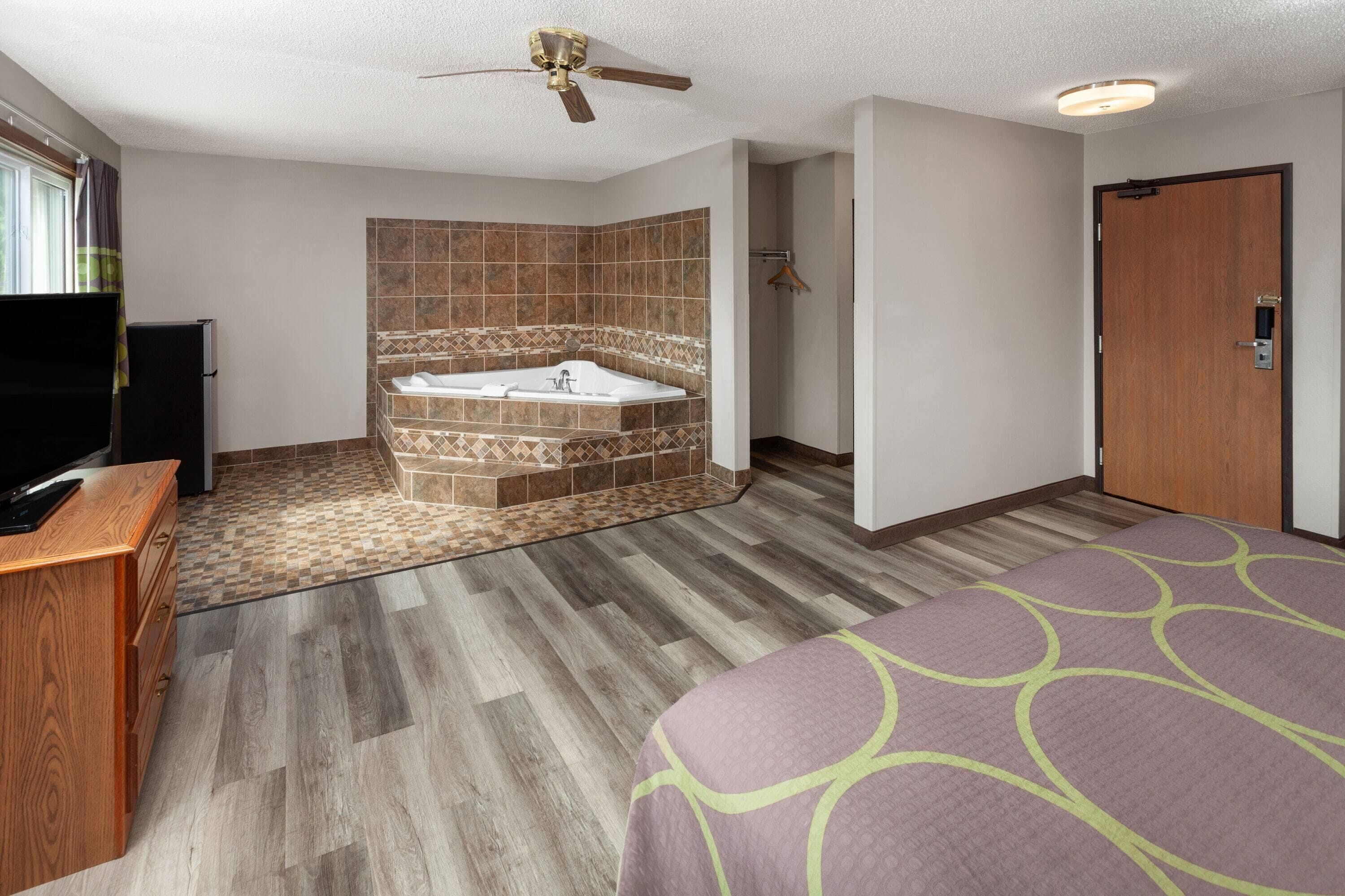 A hotel room with a wooden floor, a large bed with a green and purple bedspread, a wooden desk with a flat-screen TV, a bathroom with a bathtub and shower, and a ceiling fan.