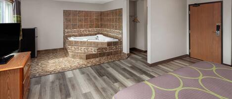 A hotel room with a wooden floor, a large bed with a green and purple bedspread, a wooden desk with a flat-screen TV, a bathroom with a bathtub and shower, and a ceiling fan.