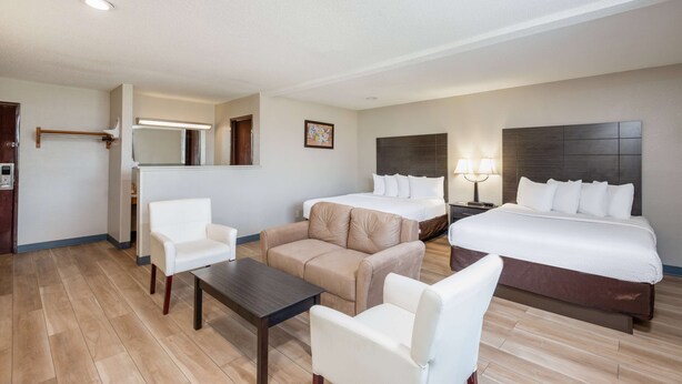 A hotel room with a bed, two white armchairs, a beige sofa, a small table, and a nightstand with lamps.