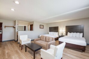 A hotel room with a bed, two white armchairs, a beige sofa, a small table, and a nightstand with lamps.