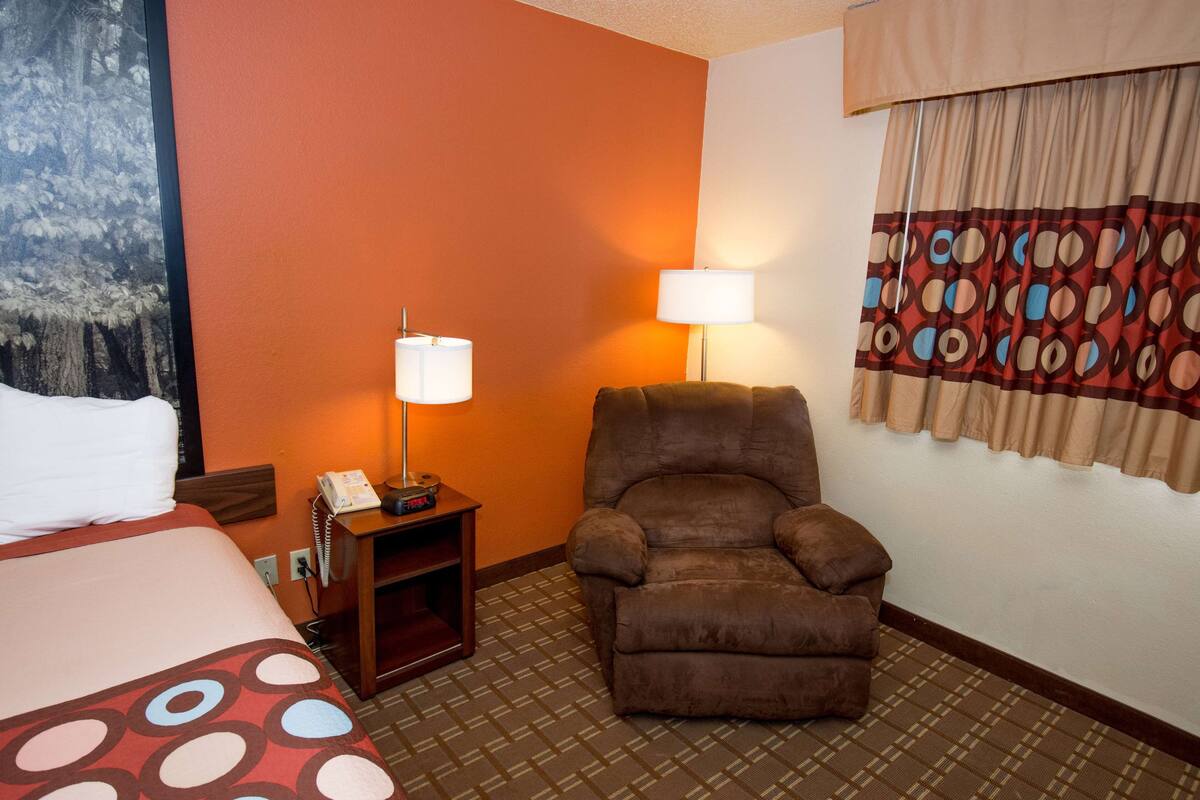 Una habitación de hotel con una cama, una silla, una mesita de noche con una lámpara y un cuadro en la pared.