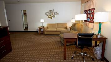 A hotel room with a patterned carpet, a beige sofa, a desk with a chair, two bedside lamps, and a decorative wall piece.