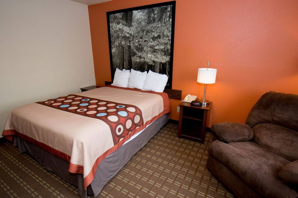 Una habitación de hotel con una cama, una mesita de noche con lámpara, un sillón marrón y un ventanal con vistas a un bosque.