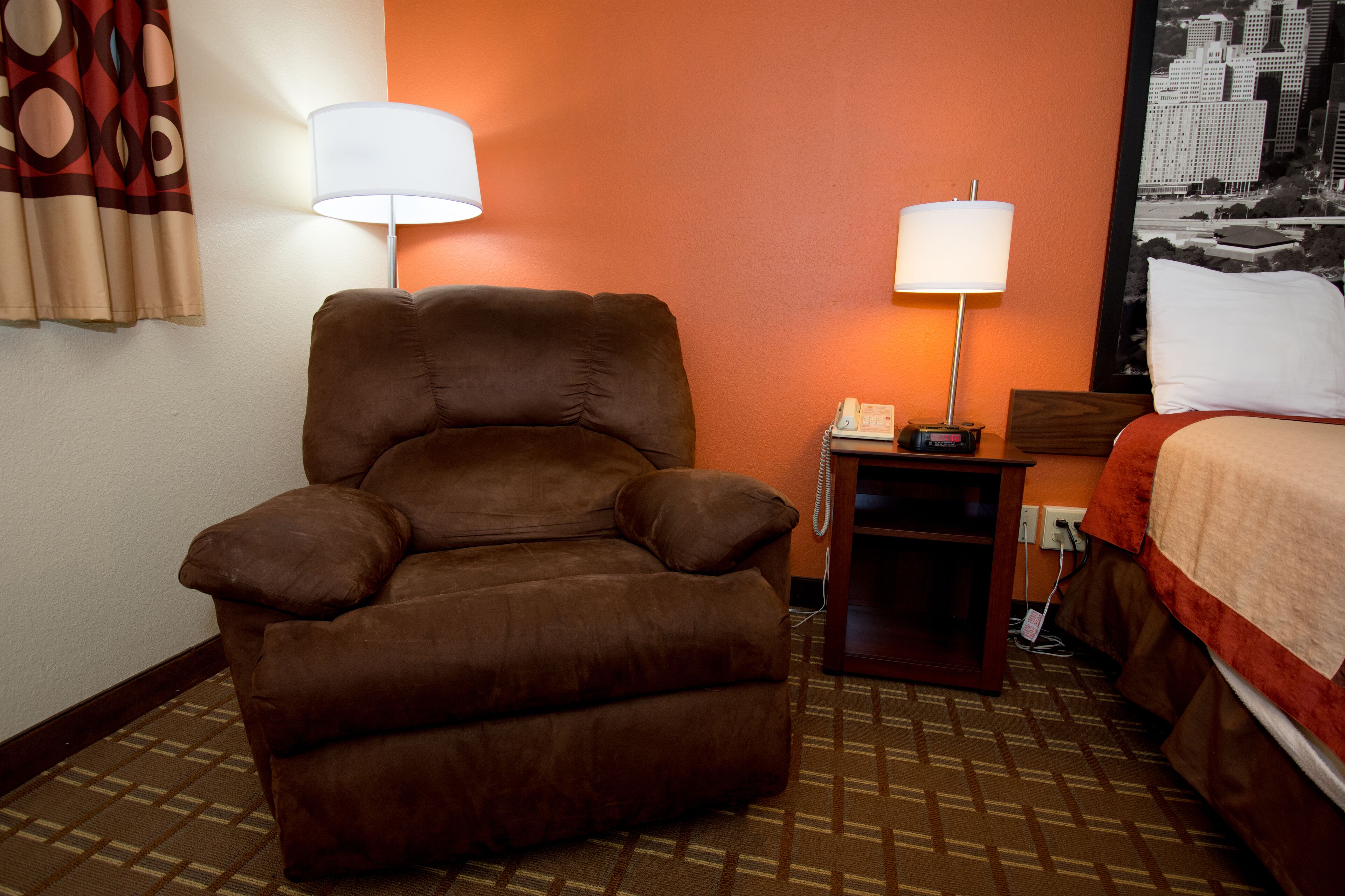 A hotel room with a brown leather armchair, two bedside lamps, a small table with a phone, and a bed with a red and white bedspread.