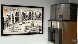 Ein gerahmtes Willkommensschild mit dem Schriftzug "Altoona" und einer Illustration eines Gebäudes, neben einem Edelstahlkühlschrank.