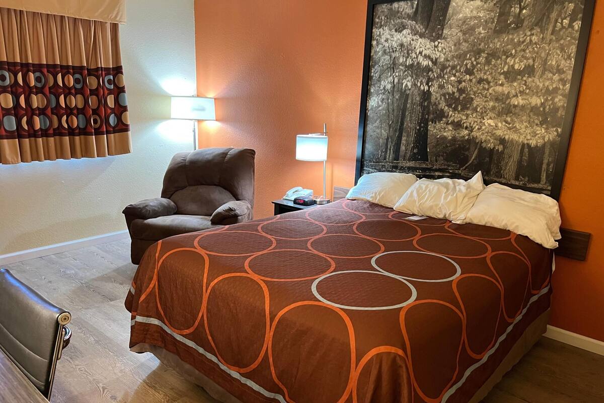 Una habitación de hotel con una cama grande, una silla, una mesita de noche con lámpara y un cuadro en la pared.