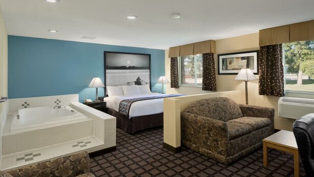 A hotel room with a large bed, a separate sofa, a small table, a desk, a desk lamp, a window with curtains, a painting on the wall, a bathtub, and a shower. - Fairview Inn & Suites Whitewater (Whitewater)