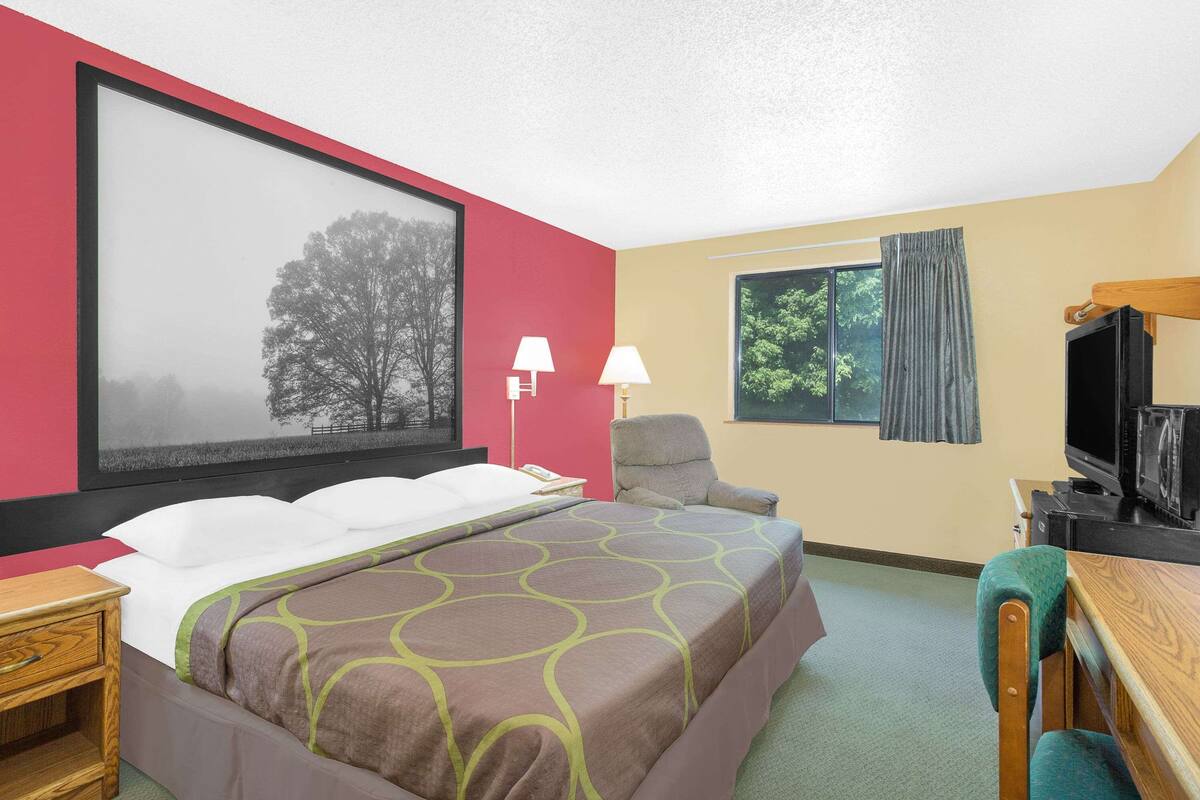 Ein Hotelzimmer mit einem großen Bett, einem Schreibtisch, einem Sessel, einem Fernseher und einem Fenster mit Vorhängen.