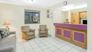 Een kamer met een bank, twee fauteuils, een bijzettafeltje met een lamp, een keuken met houten kastjes en een klok aan de muur.