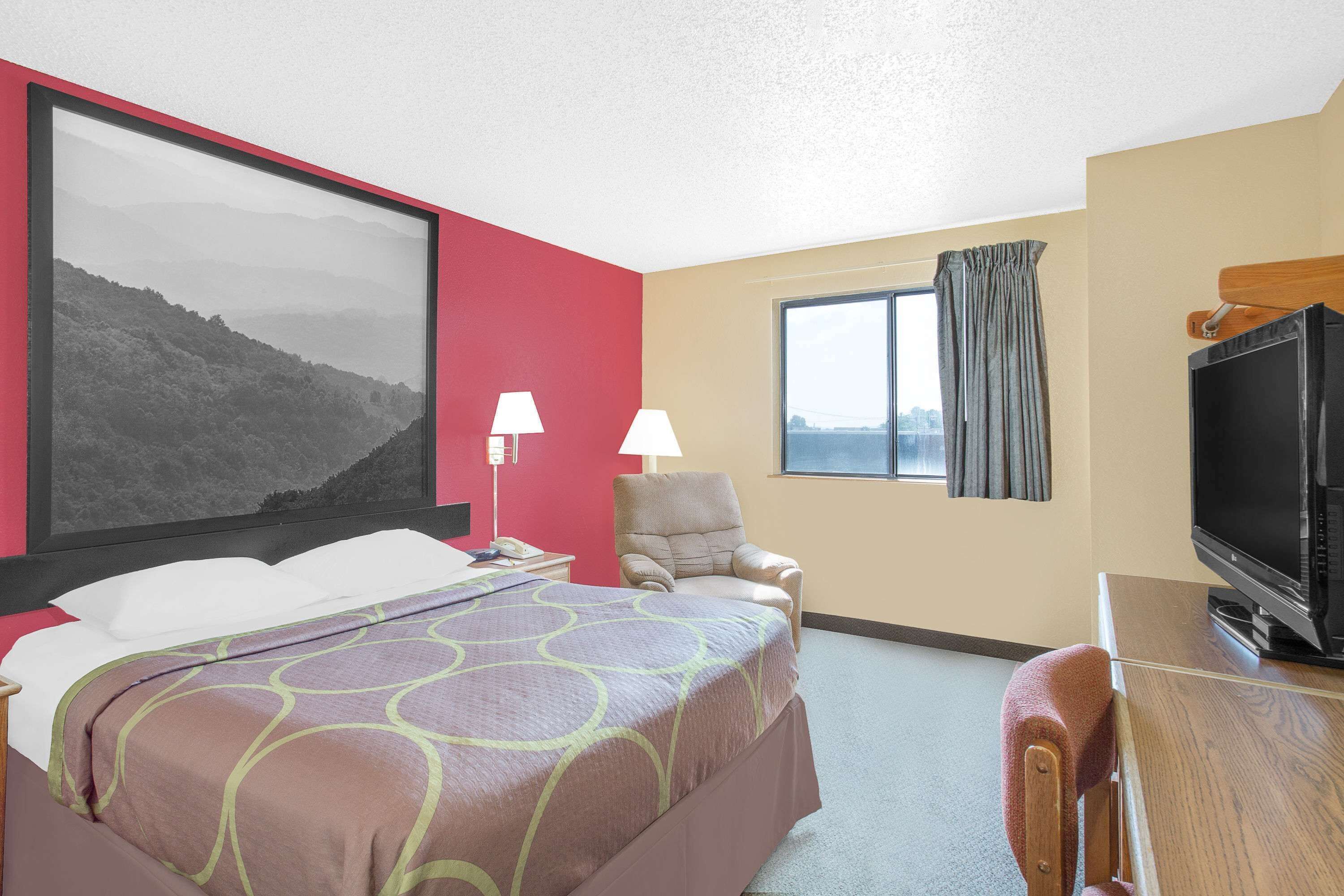 Une chambre d’hôtel avec un grand lit, une chaise, un bureau avec une télévision, une fenêtre avec des rideaux et un tableau au mur.