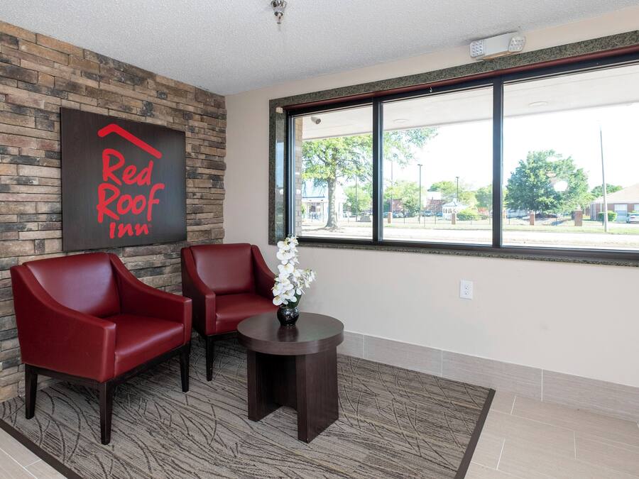 A Red Roof Inn lobby with a stone wall, two red chairs, a small table with a vase of flowers, and a large window with a view of the street.