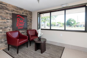 Die Lobby eines Red Roof Inn mit einer Steinwand, zwei roten Stühlen, einem kleinen Tisch mit Blumenvase und einem großen Fenster mit Blick auf die Straße.
