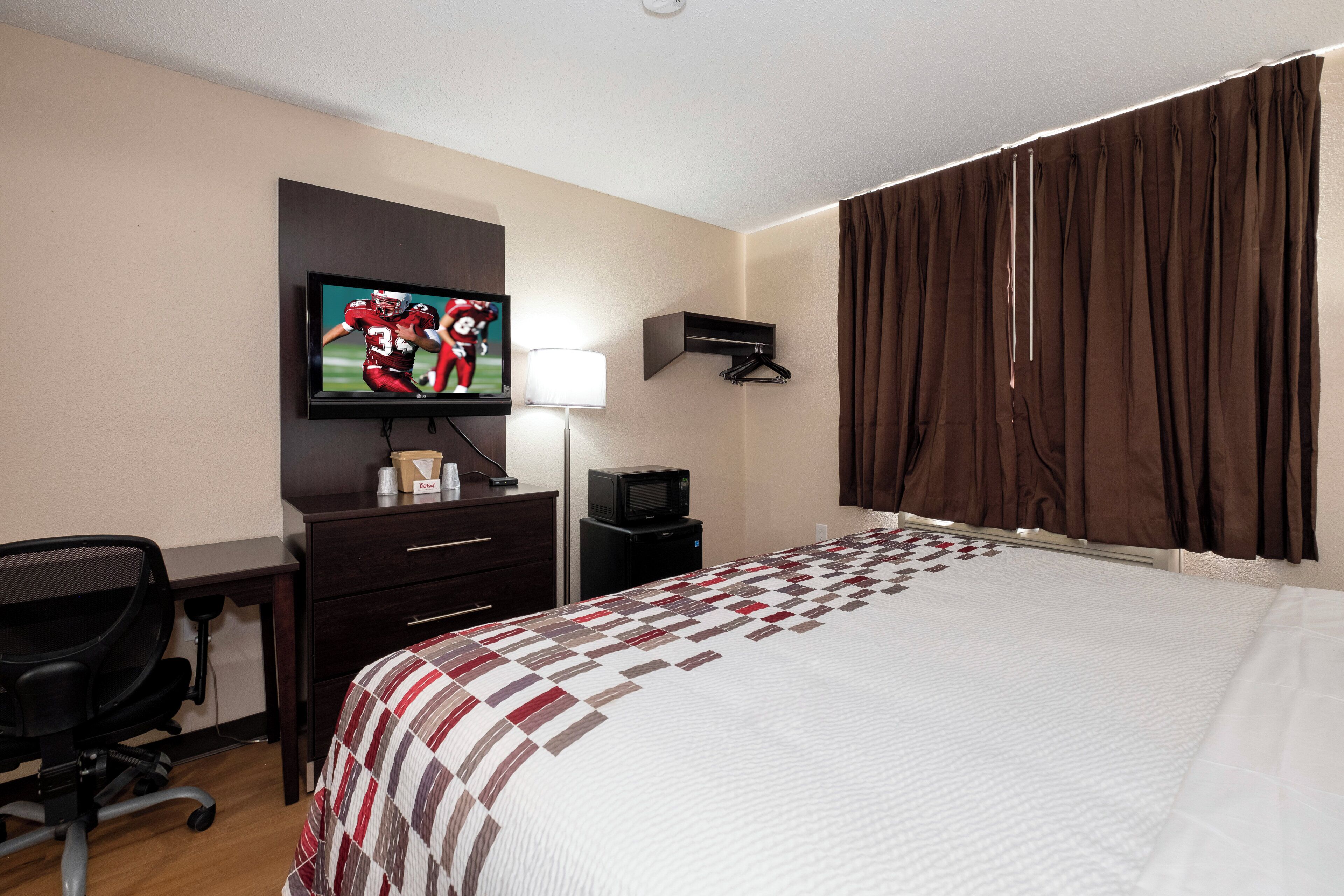 A hotel room with a bed, a desk with a chair, a television, a lamp, a coat rack, and a window with curtains.