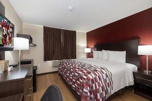 Habitación de hotel con una cama grande, un escritorio con lámpara, una silla y un televisor en la pared.