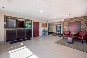 A hotel lobby with a reception desk, a red door, a wall-mounted sign, and a seating area with two red chairs.
