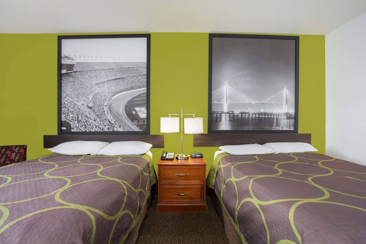 Quarto de hotel com duas camas, uma mesa de cabeceira com abajur e duas imagens emolduradas na parede.
