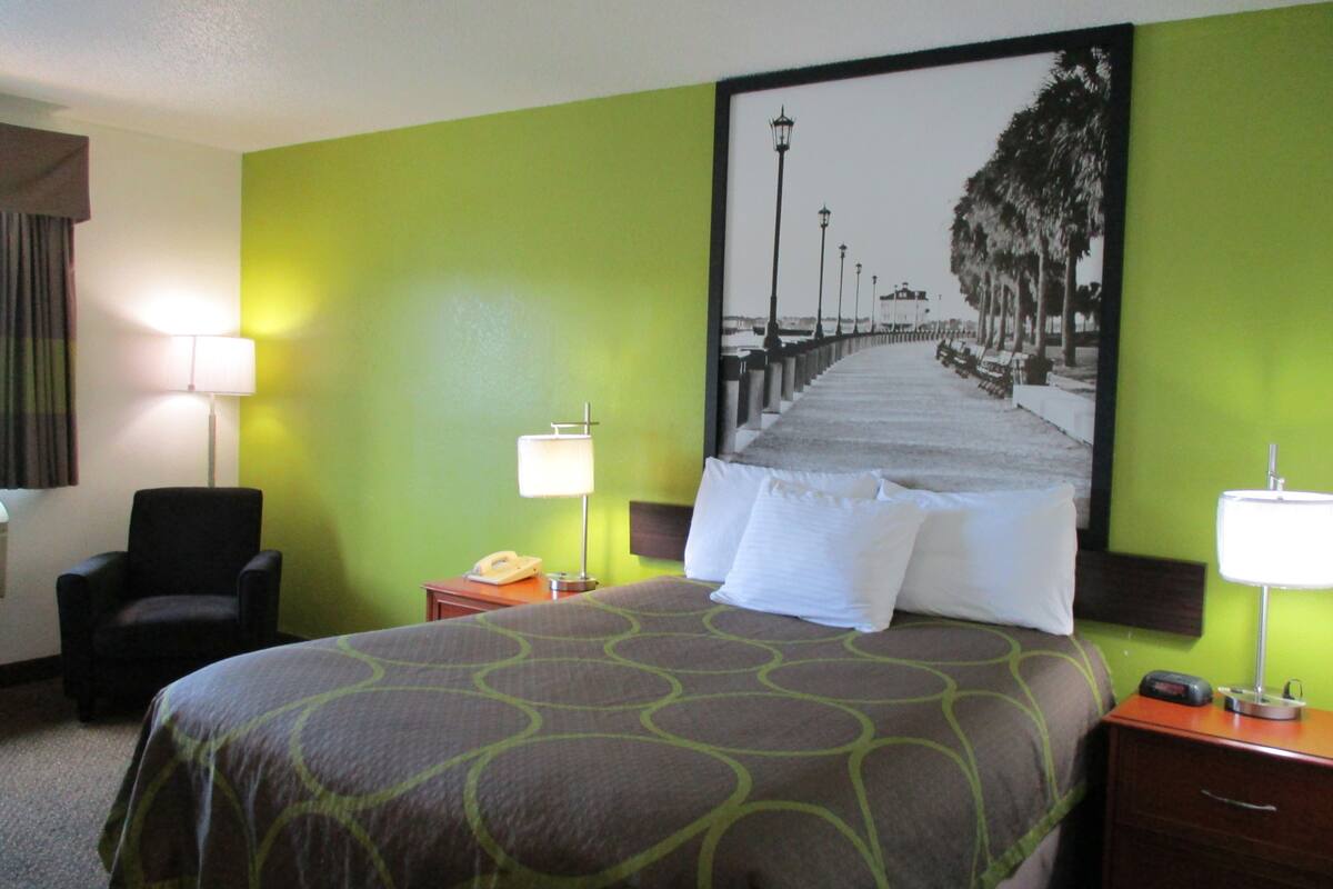 Quarto de hotel com cama, poltrona, mesa de cabeceira, abajur e parede com uma pintura.