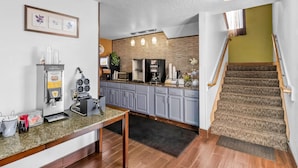 Una cocina moderna con microondas integrado, cafetera y refrigerador. Hay un comedor con mesa y sillas, y una escalera que lleva a un nivel superior.
