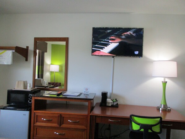 A hotel room with a desk, a chair, a television mounted on the wall, a lamp, and a microwave.