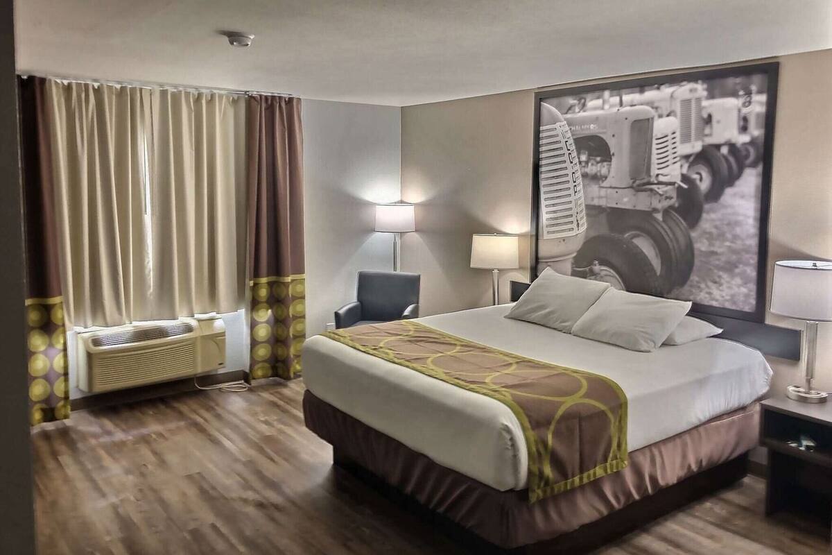 Una habitación de hotel con una cama grande, un escritorio, una silla, una lámpara, una ventana con cortinas y un aire acondicionado instalado en la pared.