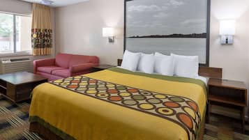 A hotel room with a bed, a red armchair, a large window with curtains, a wall-mounted lamp, and a painting of a lake.