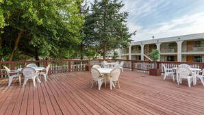 Una terraza de madera con mobiliario exterior blanco, rodeada por una valla de madera y árboles.