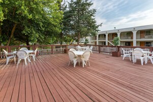 Una terraza de madera con mobiliario exterior blanco, rodeada por una valla de madera y árboles.