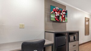 A hotel room with a kitchenette, a flat-screen TV mounted on the wall, a gray chair with a metal frame, and a wooden floor. - Red Roof Inn Suffolk (Suffolk)