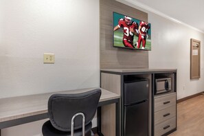 A hotel room with a kitchenette, a flat-screen TV mounted on the wall, a gray chair with a metal frame, and a wooden floor.