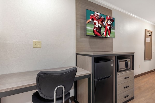 A hotel room with a kitchenette, a flat-screen TV mounted on the wall, a gray chair with a metal frame, and a wooden floor.