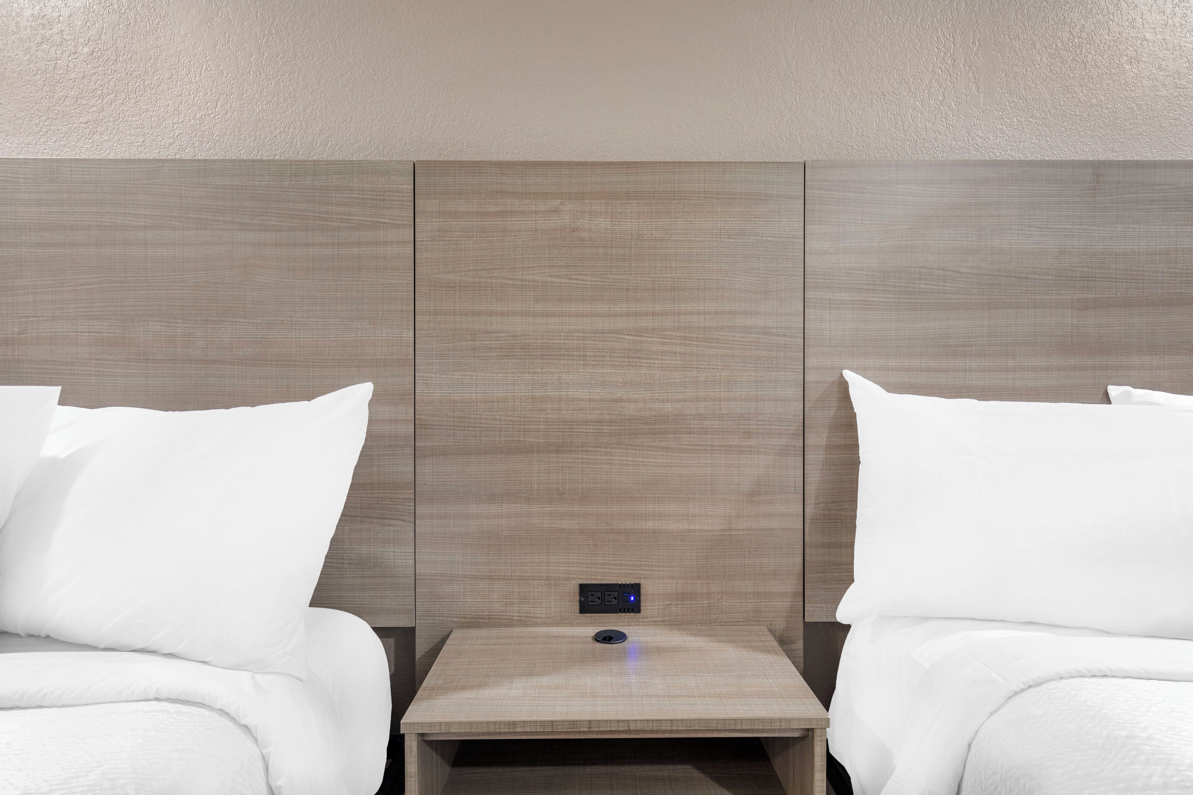 Habitación de hotel con dos camas, una mesita de noche con toma de corriente y una pared con textura.